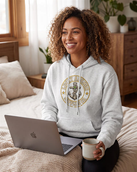 Smiling woman in a light grey Lucky Anchor crest hoodie working on a laptop in bed