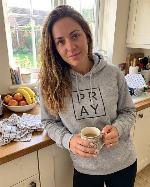 Woman in a grey PRAY hoodie holding a mug in a kitchen