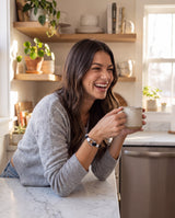 Smiling brunette woman with a coffee mug in a sunny kitchen wearing the black and white nautical paracord shackle bracelet