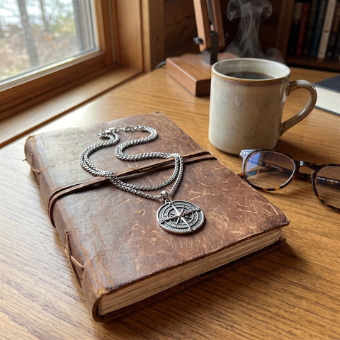 Layered compass pendant necklace set on a leather journal beside a mug and glasses