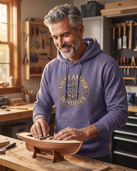 Silver-haired man in a dusty purple Lucky Anchor crest hoodie sanding a model sailboat in a workshop