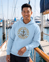 Smiling man in a light blue Lucky Anchor crest hoodie leaning on a sailboat in a marina