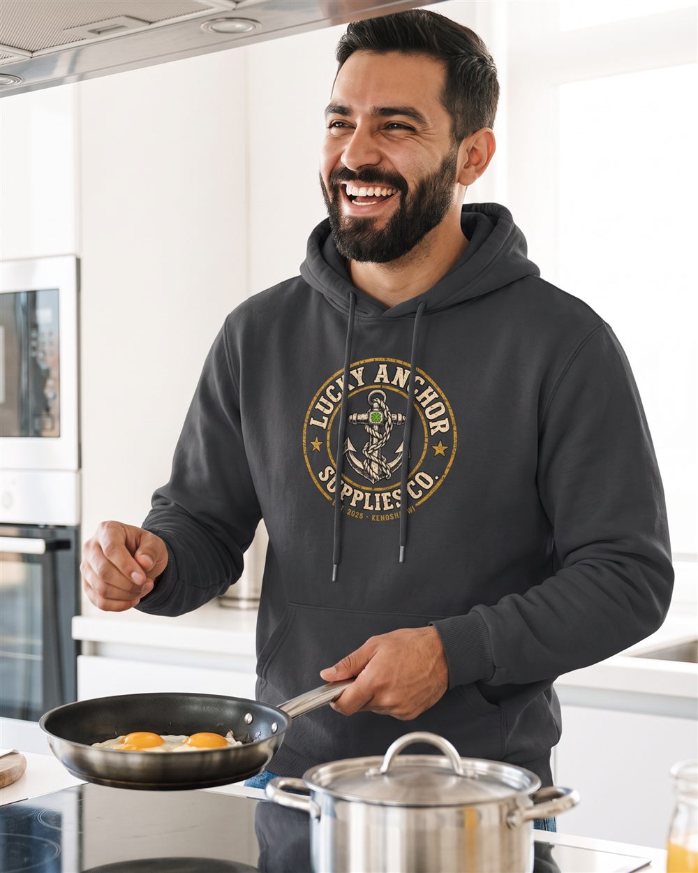 Smiling bearded man in a black Lucky Anchor crest hoodie with white drawstrings cooking in a bright kitchen