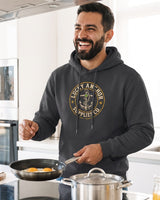 Smiling bearded man in a black Lucky Anchor crest hoodie with white drawstrings cooking in a bright kitchen