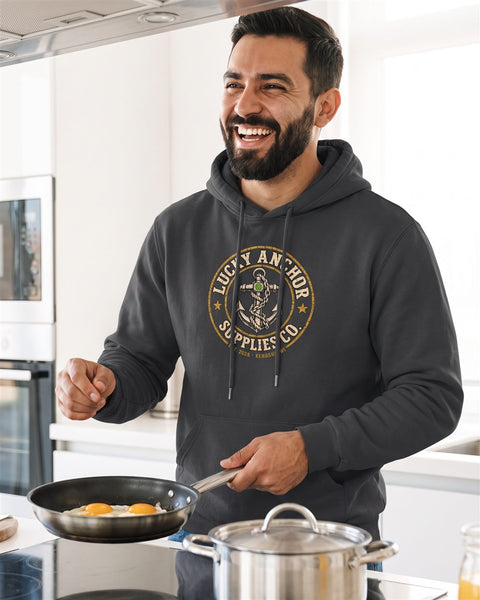 Smiling bearded man in a black Lucky Anchor crest hoodie with white drawstrings cooking in a bright kitchen