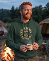 Red-haired man in a forest green Lucky Anchor crest hoodie holding a mug by a campfire