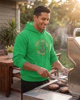 Smiling man in a green Lucky Anchor crest hoodie grilling burgers on a backyard patio