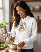Smiling woman in a white Lucky Anchor crest hoodie pouring tea in a farmhouse kitchen