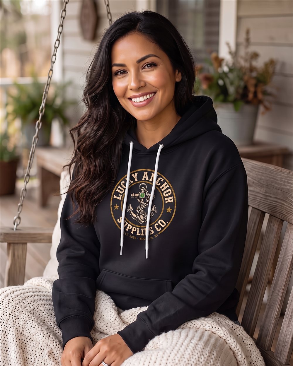 Smiling woman in a black Lucky Anchor crest hoodie wrapped in a blanket on a porch swing