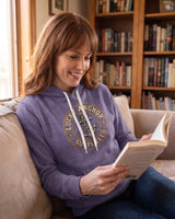 Woman in a dusty purple Lucky Anchor crest hoodie reading on a living room sofa