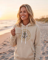 Smiling woman in a sand Lucky Anchor crest hoodie holding a coffee on a beach at sunrise