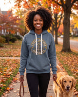 Smiling woman in a slate blue Lucky Anchor crest hoodie walking a dog on an autumn sidewalk